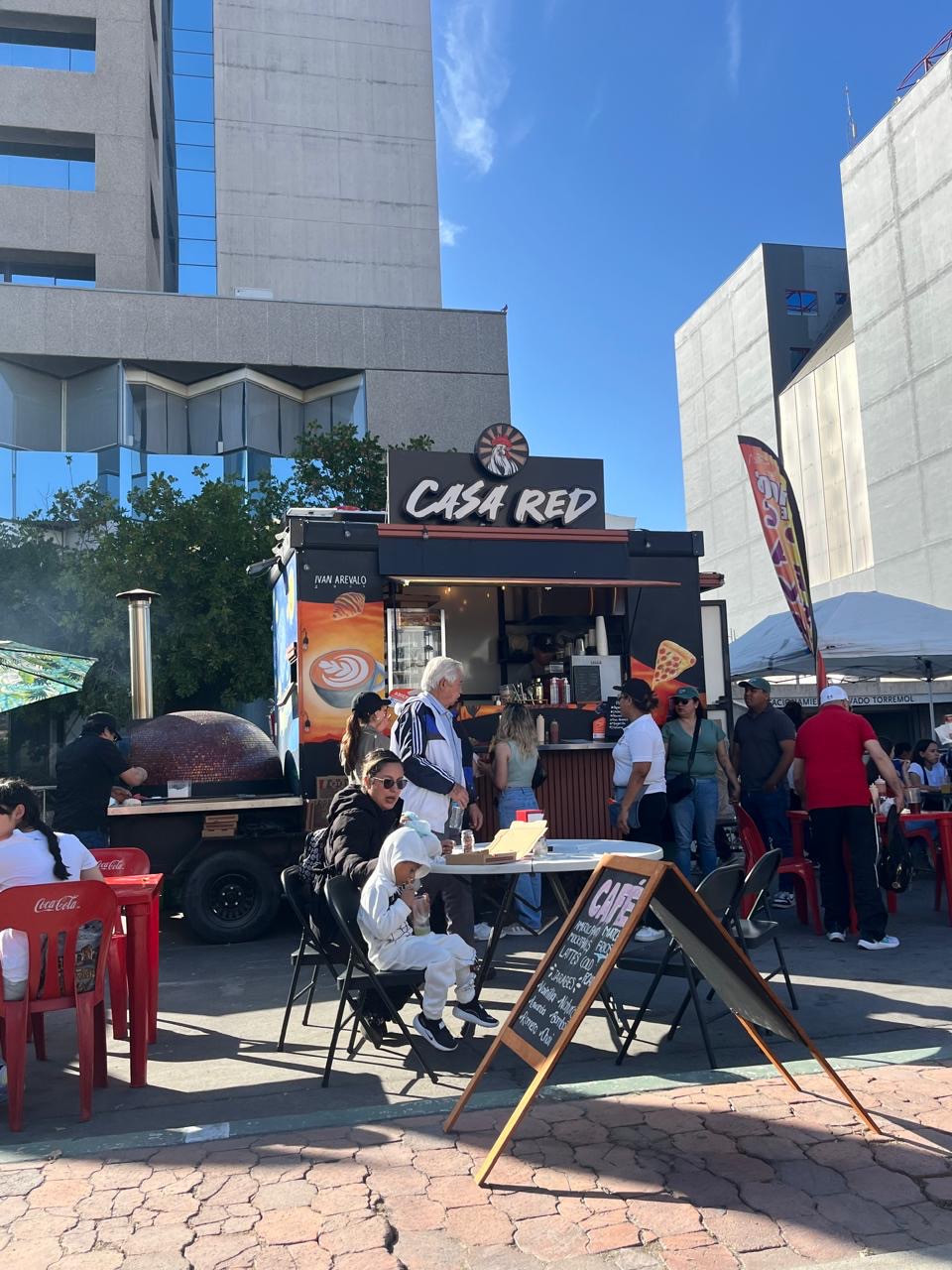 Carrito Casa Red en un evento de día en una plaza, con mesas, clientes, horno de pizza y edificios al fondo.
