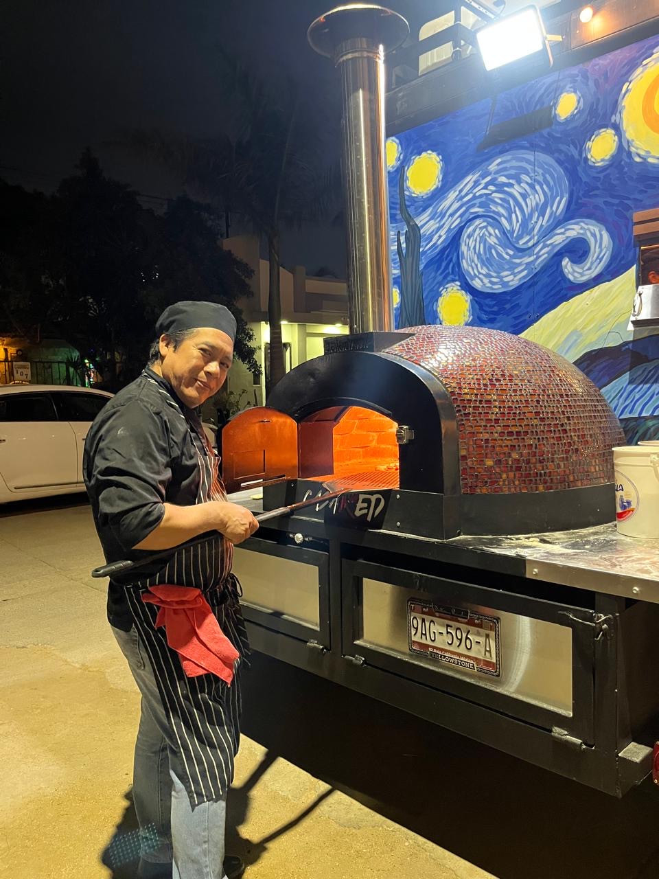 Cocinero junto al horno de pizza encendido durante la noche, con la pala del horno y el mural azul del carrito.