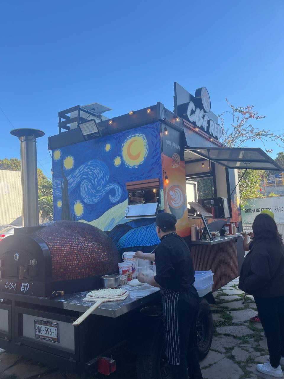 Preparación de una pizza junto al horno del carrito Casa Red durante el día, con el mural azul y clientes junto al mostrador.